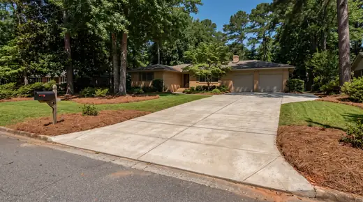 Wide concrete driveway viewed from the street toward the garage at a buff-brick ranch home in Savannah, GA, in clear late-morning sun.